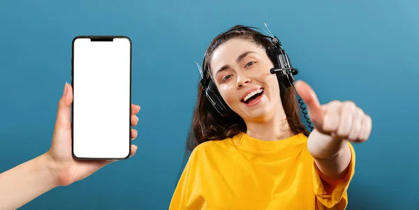 Online courses. Portrait of a young smiling woman wearing headphones, giving a thumbs-up. Female hand holding a cellphone with mock up. Blue background. Concept of online learning