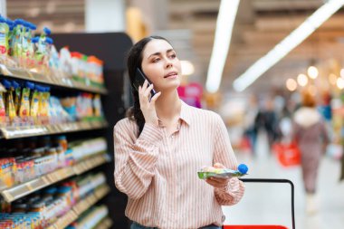 Gülümseyen genç kadının portresi el arabası, ürünler ve cep telefonuyla yapılan aramaları barındırıyor. Arkaplanda yiyecek olan raflar. Marketten alışveriş kavramı.