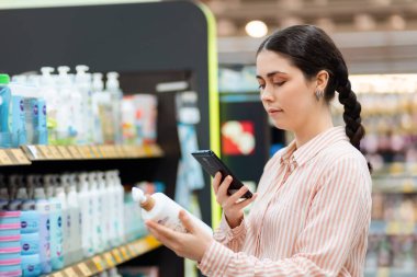 Kafkasyalı genç bir kadının akıllı telefon tutarken ve kozmetik şişesinde soru cevap kodlarını tararken yan görüntüsü. Modern alışveriş kavramı.