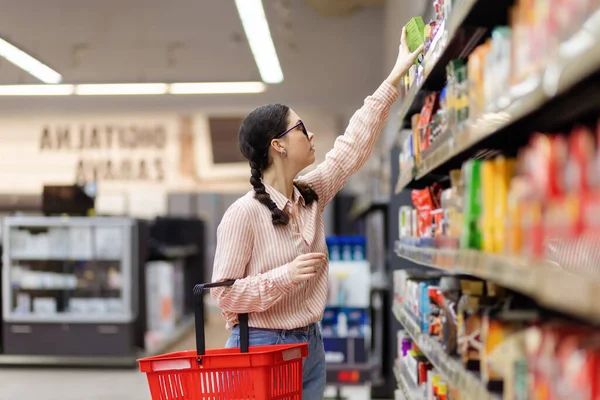 Visit grocery store. Side view of young pretty caucasian woman holds cart and reaches hand to top shelf. Concept of shopping and consumerism.