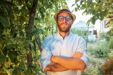 Hasır şapkalı ve gözlüklü kendine güvenen beyaz adamın portresi bahçede pozlar veriyor, çapraz eller. Organik bahçe ve tarım kavramı..