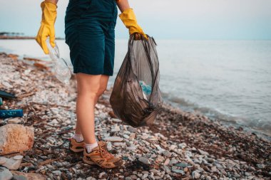 Dünya günü. Bir gönüllü, içinde çöp olan plastik bir poşet tutuyor ve sahilde poz veriyor. Ayaklara yakın çekim. Okyanus kıyısındaki çöpleri temizleme kavramı.
