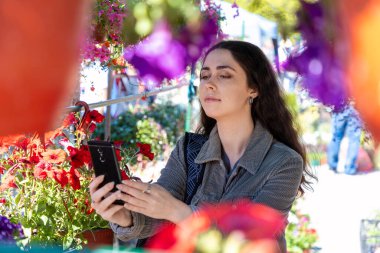 Beyaz tenli güzel bir kadın çiçek saksılarının fotoğrafını çekiyor. Çiçekçi bitkileri seçer..