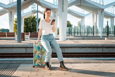 Bavulunun üzerinde akıllı telefon kullanan beyaz bir kadın. Tren istasyonu arka planda. Seyahat ve tatil kavramı.