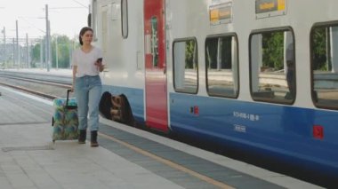 Gülümseyen güzel beyaz kadın akıllı telefon kullanıyor ve bavulunu istasyonda tutuyor. Kalkış treni arka planda. Seyahat ve varış.