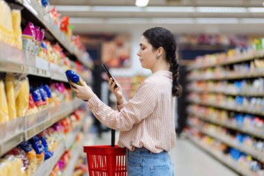 Side view of caucasian woman scan qr code of product using smartphone. Shelves with food in background. Concept of shopping and consumerism. clipart