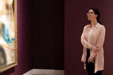 Genç, güzel, beyaz bir kadın sanat sergisine bakıyor. Arka planda insanlar ve şaheserler var. Uzayı kopyala Müze Günü Konsepti.