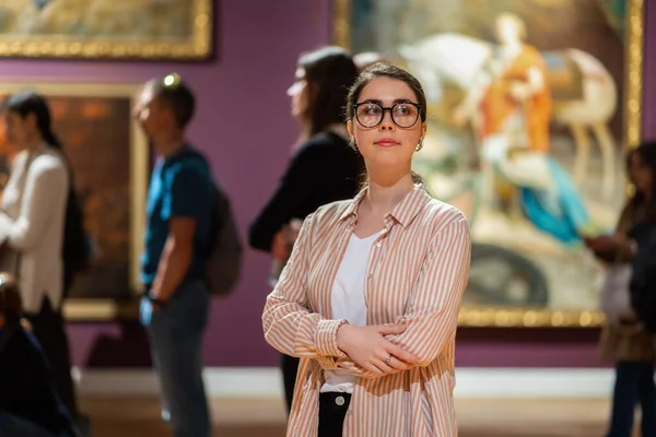 stock image Young Caucasian woman wearing glasses and looking at exhibition. People and masterpieces in background. Concept of Museum Day.