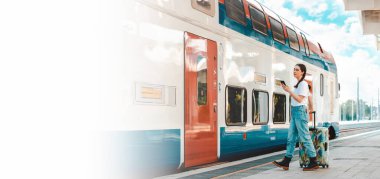 Web seyahat afişi ve maket. Ayrılan genç ve güzel bir kadın bavulunu tutar ve cep telefonu kullanır. Tren istasyonu arka planda. Tatil ve iş gezisi kavramı.