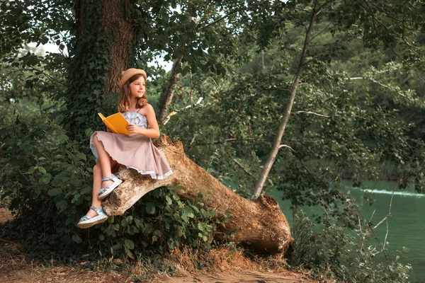 穿着裙子 头戴草帽 头戴草帽 拿着一本书坐在一根旧圆木上 梦想着一个女学生 背景是公园和河流 复制空间 课外阅读和儿童教育的概念 — 图库照片