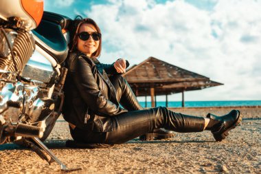 Deri motosikletli bağımsız mutlu kadın motosikletin yanında poz veriyor. Yan görüş. Arka planda okyanus ve plaj şemsiyeleri var. Motosiklet yolculuğu ve feminizm konsepti.