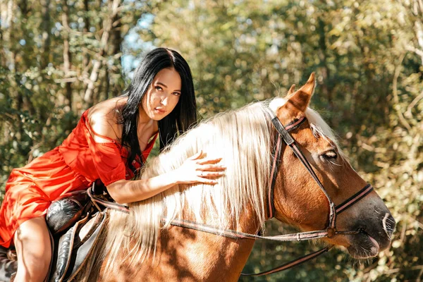一个穿着红色衣服的年轻漂亮的黑发女人骑在一匹棕色的马的前面 — 图库照片