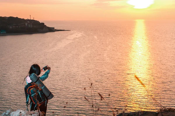 Bir kadın uçurumun kenarında duruyor ve akıllı telefonuyla günbatımının fotoğraflarını çekiyor. Arka planda, gün batımında deniz. Uzayı kopyala ve giyin. Yürüyüş ve açık hava aktiviteleri kavramı.