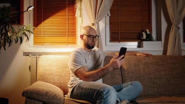 ソファーで自宅に座っているスマートフォンを使用して 若いひげを被った白人男性の肖像画 夜の雰囲気と電灯について オンラインビデオ通話の概念 — ストック動画