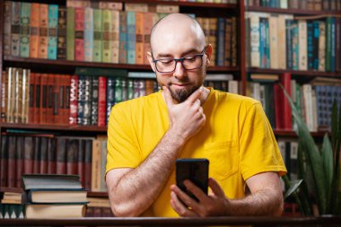 İletişim. Gözlüklü, gülümseyen kel bir adam akıllı telefon kullanır. Arka planda bir kütüphane ve kitap rafları var. Sosyal ağlar, uzaklık öğrenme ve karantina kavramı.