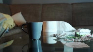 Close up of glass table with medicines and mess. Tracking left shot to suffering Caucasian woman lies on sofa covered with blanket and watching tv. Flu and depression.