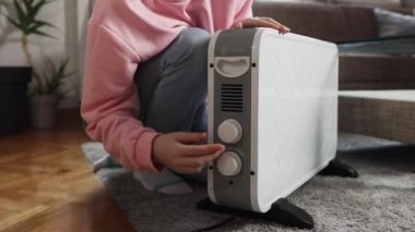 Pembe kazaklı ve kotlu bir kadın elektrikli ısıtıcıyı açıyor ve kendini ısıtıyor. Yakın plan. Gaz ve ekonomik ısıtma sisteminin reddi kavramı