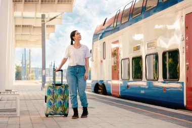 Bagajını tutan ve tren istasyonundan kalkmayı bekleyen genç bir kadın. Arka planda renkli tren var. Seyahat ve tatil kavramı.