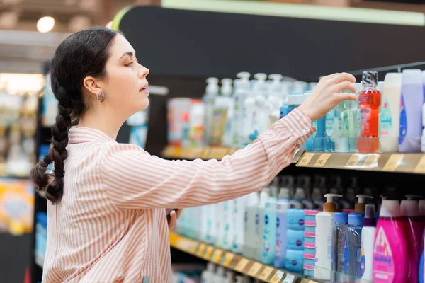 Portrait of Caucasian young woman takes shampoo bottle from shelf. Choice of care cosmetics in supermarket. Concept of shopping and purchase.