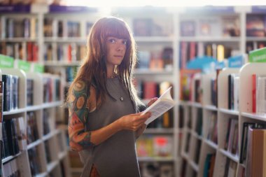 Dövmeli beyaz bir kadın kitap tutuyor ve kitapçıda poz verirken kameraya bakıyor. Eğitim ve alışveriş kavramı.
