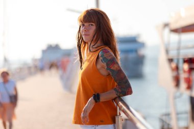 İskeleye dayanan ve kameraya bakan dövmeli genç bir kadın. Arka planda iskele. Özgürlük ve yaz tatili.