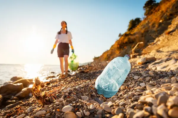 Defocused view of woman volunteer holds full plastic bag of trash and walking down of pebble wild beach. Close up of plastic bottle in foreground. Concept of protection ocean and Earth Day.