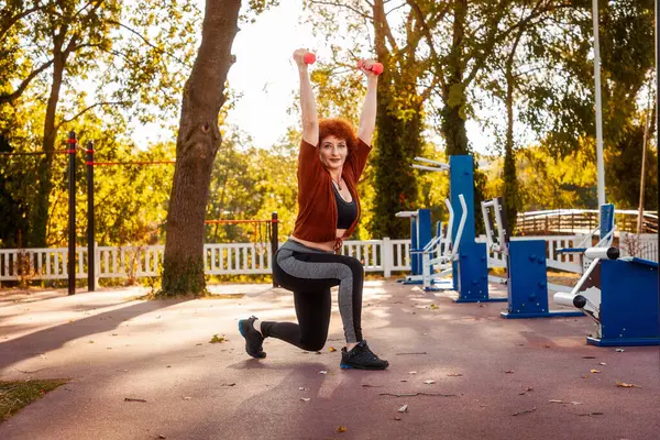 Adult Caucasian strength woman posing with dumbbells squatting in park on street and looking in camera. Copy space. Healthy lifestyle.