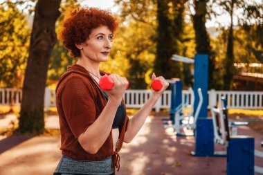 Yetişkin güçlü beyaz kadın portresi sokakta parkta spor yapıyor. Sporcu elinde dambıllarla çömelir. Sağlıklı yaşam tarzı.