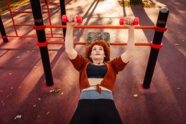 Beyaz, orta yaşlı, güçlü bir kadının şehir parkındaki yedek kulübesinde Fransızca göğüs presi yaptığı görülüyor. Spor ve spor yaşam tarzı kavramı.