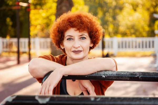 Portrait of adult Caucasian woman with makeup and ginger curly hair posing in sports park. Concept of healthy lifestyle.