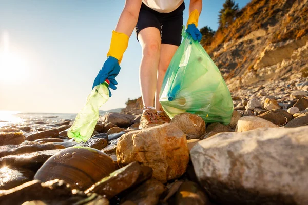 Bottom view of woman volunteer\'s hand in rubber gloves takes plastic bottle from pebble wild coast. Concept of ocean pollution.