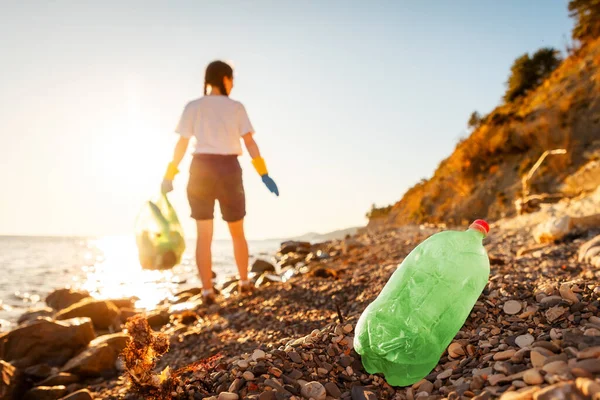 Defocused back view of woman volunteer in rubber gloves holds full plastic bag of trash. Pollution of pebble wild beach. Concept of protection ocean and Earth Day.