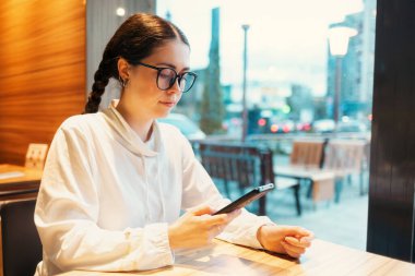 Serbest çalışıyorum. Gözlüklü genç beyaz kadın pencere kenarında oturuyor ve akıllı telefon kullanıyor. Kafelerde kablosuz internet ve iş birliği..