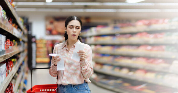 Web banner of shopping and high price. Portrait of shocked Caucasian woman holds paper receipts of purchase. In background, rows of shelves with products. Concept of inflation. Copy space.