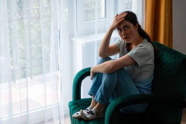 Yeşil kadife koltukta oturan üzgün, Kafkasyalı genç kadın başını tutuyor. Depresyon, psikoloji ve akıl sağlığı sorunları kavramı.