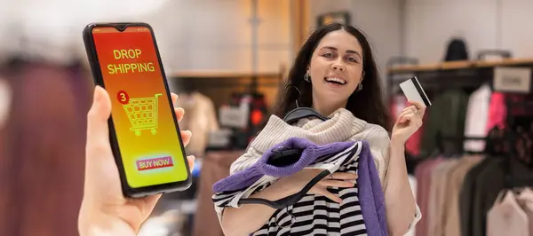 Web banner of drop shipping. Happy young woman holds hangers with clothes and bank card. Close up of female hand holding smartphone with online store at screen. Concept of online shopping and sales.