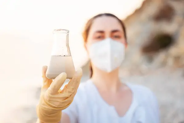 Portrait of younCaucasian woman wearing protective gloves and vest, holds research sample flask. Ecologist examines chemical dirty water test. Concept of pollution water and ecology