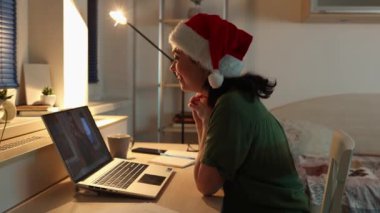 Görüntülü arama. Kafkasyalı güzel bir kadının noel şapkası takıp dizüstü bilgisayar kullanmasının yan görüntüsü. Uzaktan çalışma ve çevrimiçi iletişim kavramı.