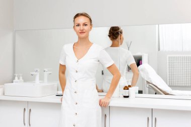 Tıp bürosunda masanın yanında poz veren gülümseyen bir kozmetik uzmanı. Profesyonel kozmetoloji kavramı.