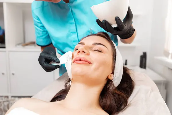 Facial beauty procedure. Beautician in latex gloves applying with spatula rejuvenation mask on the woman\'s face. Close up. Concept of professional cosmetology.