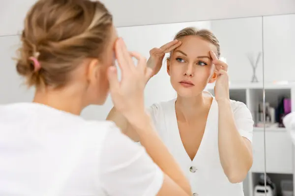 A young beautiful woman looks in the mirror and pulls the skin on her forehead. The view from behind the shoulder, at the reflection in the mirror. The concept of rejuvenation and facial wrinkles.