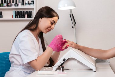 Pembe tıbbi eldivenli beyaz manikürcü müşterinin tırnaklarını parlak pembe cila ile boyuyor. Kapalı alanda. Manikür salonunda profesyonel bakım konsepti.