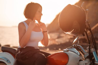 Kasklı, motosikletin yanında duran güzel, yetişkin bir kadının silueti. Günbatımı arka planda. Motosiklet yolculuğu ve feminizm kavramı.