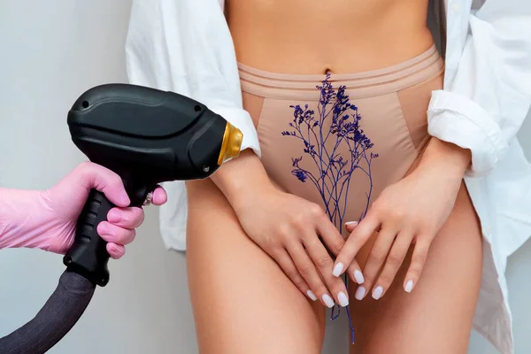 Cosmetologist holds a laser device near slim woman in beautiful underwear holding a dry herbs cover intimate area. Beige background. The concept of epilation and laser hair removal.