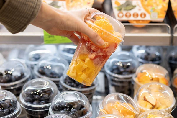 Close up of female hand holding plastic cup filled with chopped fruits. Eco-friendly, organic and healthy snack. Food in shelf in supermarket.