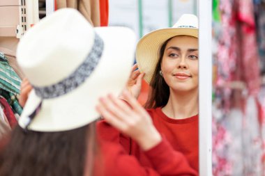 Kafkasyalı güzel bir kadın dükkanda aynanın önünde hasır şapka deniyor. Dikiz aynasından yansıyan yüz. Alışveriş konsepti.