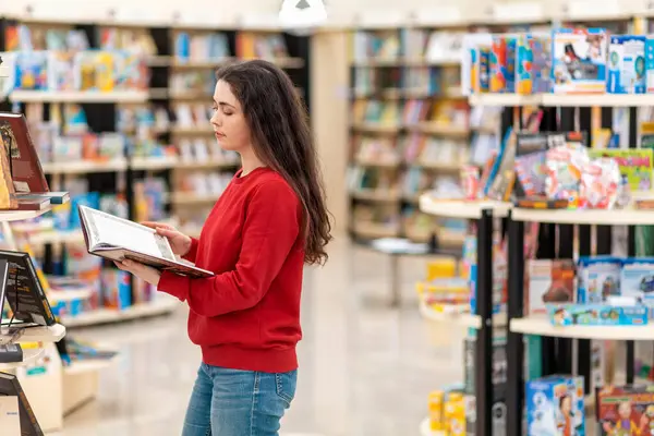 A young Caucasian woman reads a book in her hands, against the background of shelves with books. Side view. The concept of education and purchase of books.