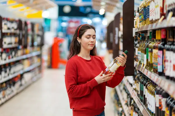 A young Caucasian woman examines a bottle of white wine. In the background, the store shelves are blurred. The concept of buying alcohol in a store.