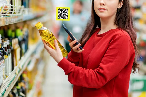 A young Caucasian woman in a red sweatshirt scans the qr code on a bottle of oil using her smartphone. Qr code icon above the product. In the background is a supermarket. Close up. Concept of modern technologies and shopping.