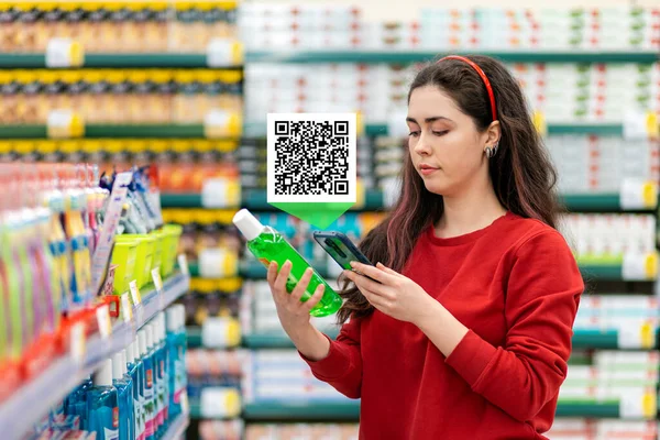 A young Caucasian woman scans the qr code on a shampoo bottle using her smartphone. Qr code icon above the product. In the background is a supermarket. Concept of modern technologies and shopping.